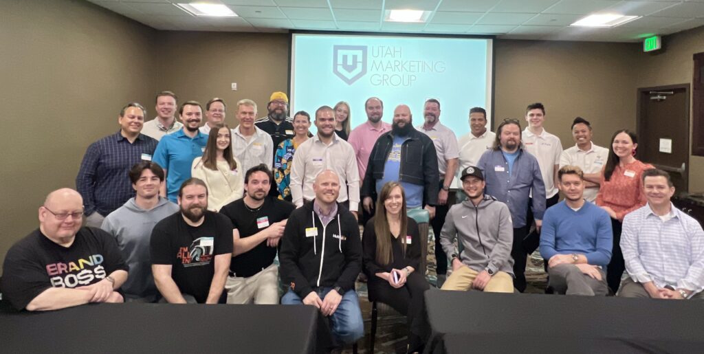 Members of the Utah Marketing Group standing and sitting in front of the UMG logo projected on the wall.