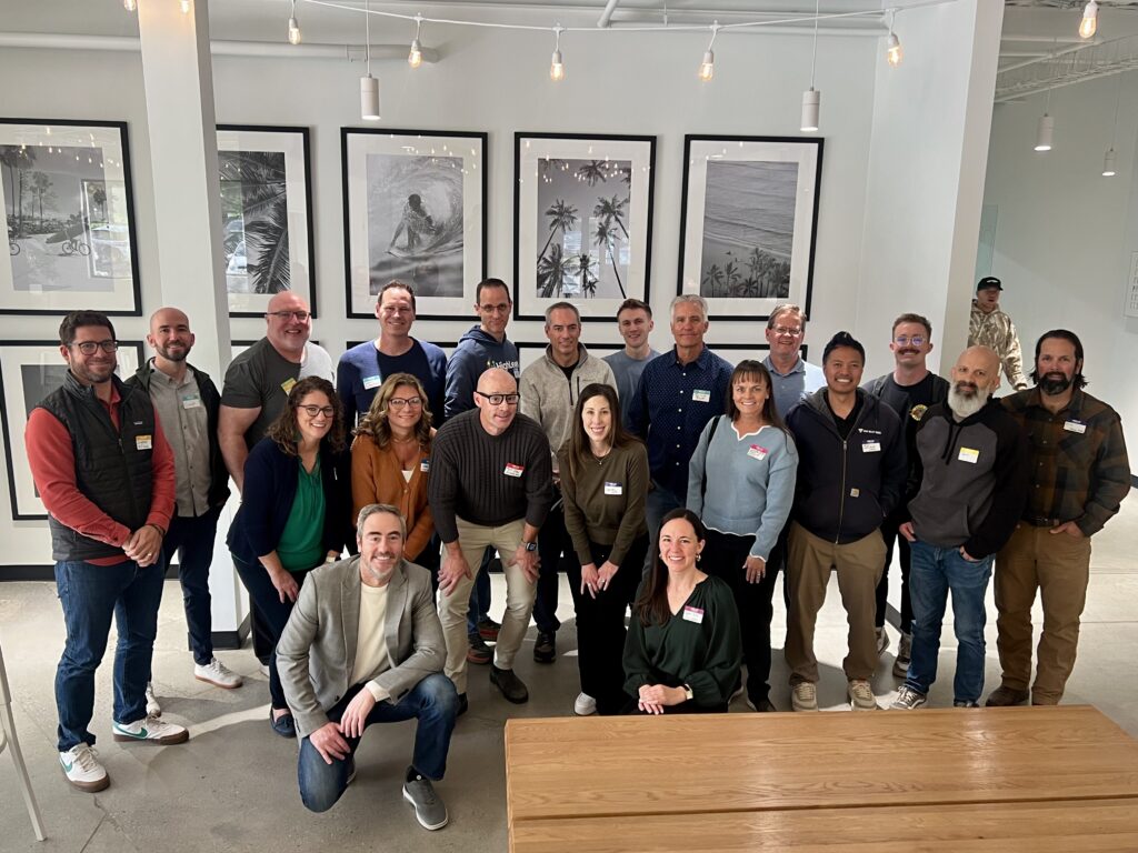 A large group of around 20 people posing together in a well-lit room. The group is smiling and dressed in casual to business-casual attire for a Utah Marketing Group meeting.