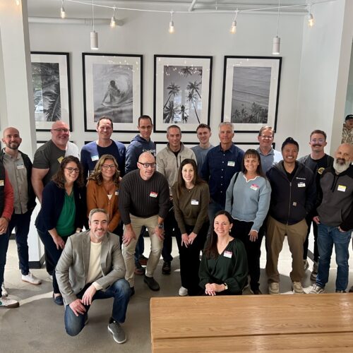 A large group of around 20 people posing together in a well-lit room. The group is smiling and dressed in casual to business-casual attire for a Utah Marketing Group meeting.