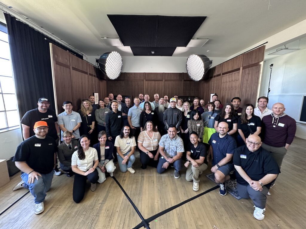 A large group of around 45 people posing together in a well-lit room with wood-paneled walls and two large studio lights overhead. The group is smiling and dressed in casual to business-casual attire for a Utah Marketing Group meeting.