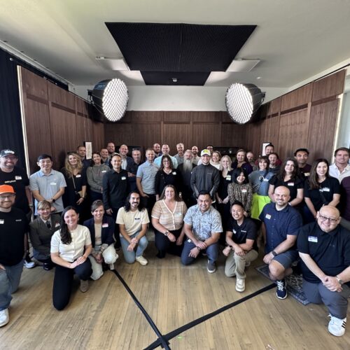 A large group of around 45 people posing together in a well-lit room with wood-paneled walls and two large studio lights overhead. The group is smiling and dressed in casual to business-casual attire for a Utah Marketing Group meeting.