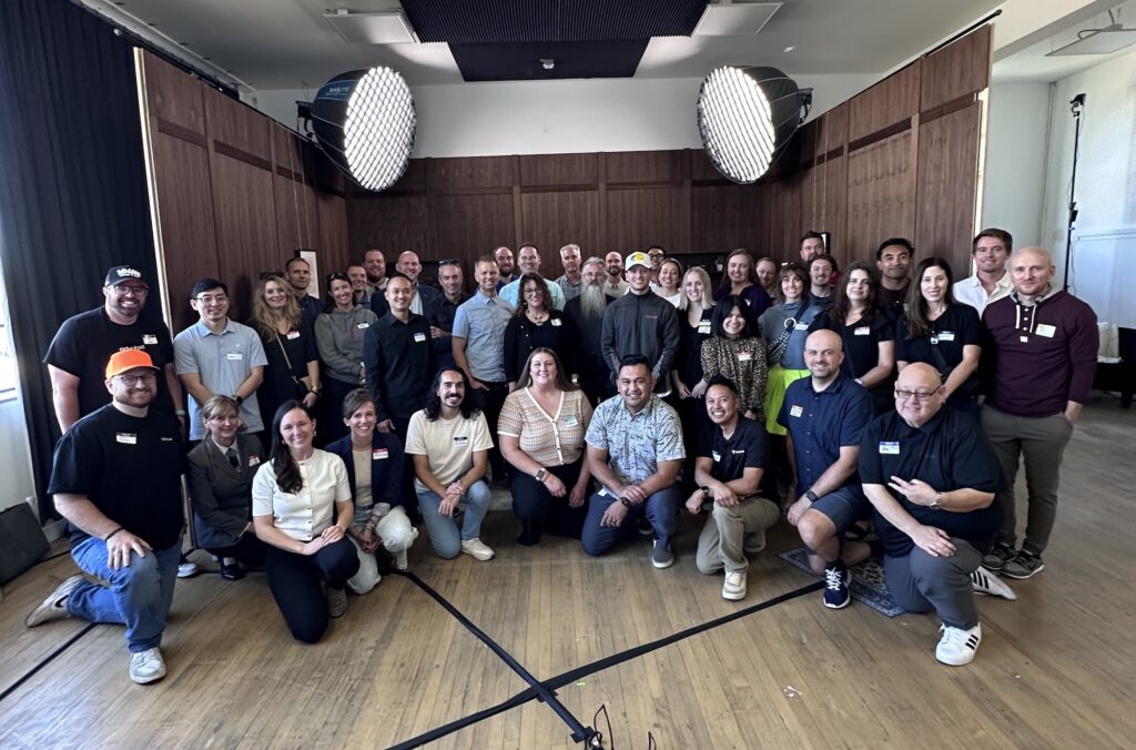 A large group of around 45 people posing together in a well-lit room with wood-paneled walls and two large studio lights overhead. The group is smiling and dressed in casual to business-casual attire for a Utah Marketing Group meeting.
