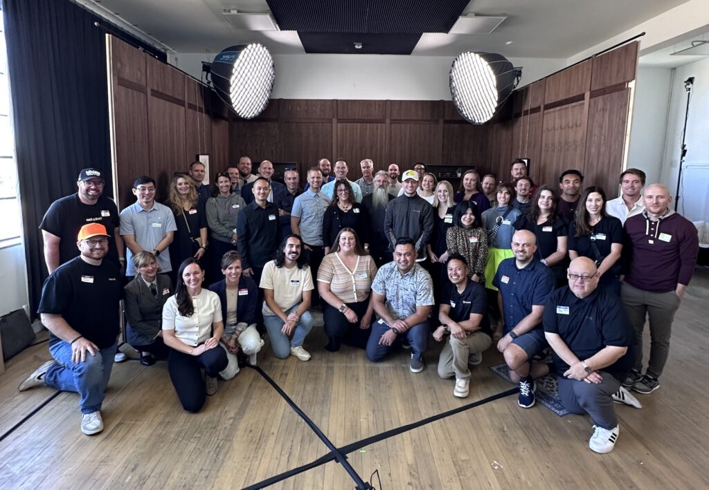 A large group of around 45 people posing together in a well-lit room with wood-paneled walls and two large studio lights overhead. The group is smiling and dressed in casual to business-casual attire for a Utah Marketing Group meeting.