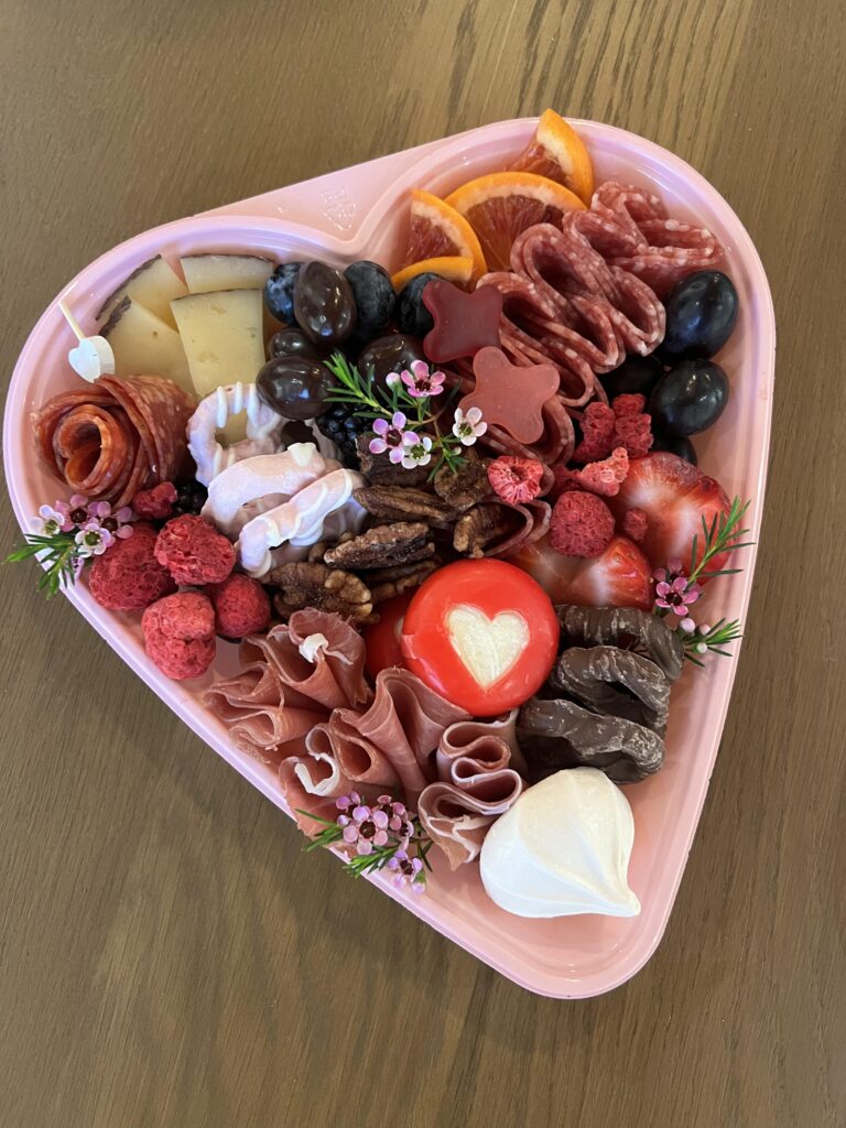 A heart-shaped charcuterie box with a mix of meats, cheeses, fruits, candies, and tiny flowers.