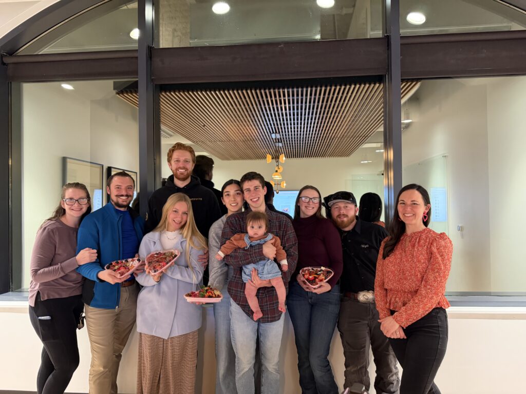 A group of 11 people standing in front of a table, holding heart-shaped charcuterie boxes they made at a class.