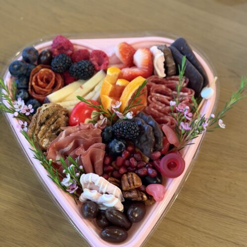 A heart-shaped charcuterie box with a mix of meats, cheeses, fruits, candies, and tiny flowers.