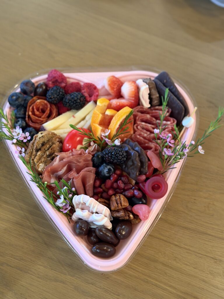 A heart-shaped charcuterie box with a mix of meats, cheeses, fruits, candies, and tiny flowers.