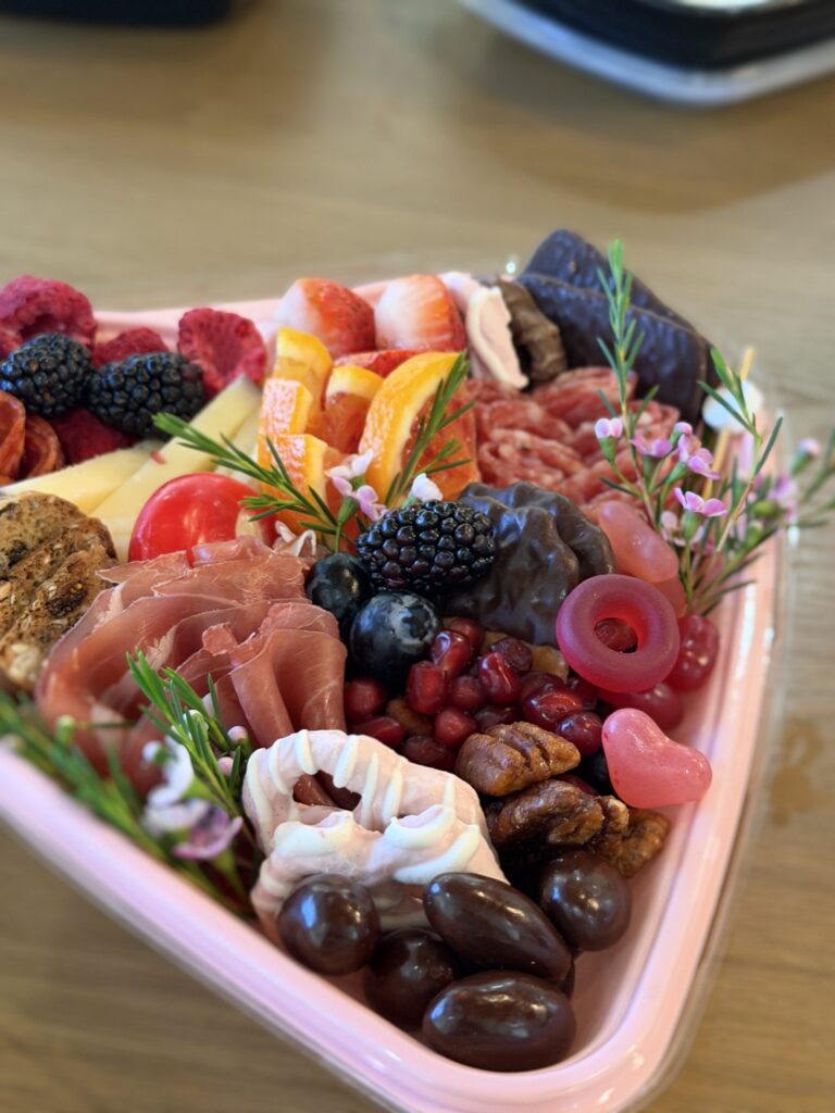 A heart-shaped charcuterie box with a mix of meats, cheeses, fruits, candies, and tiny flowers.