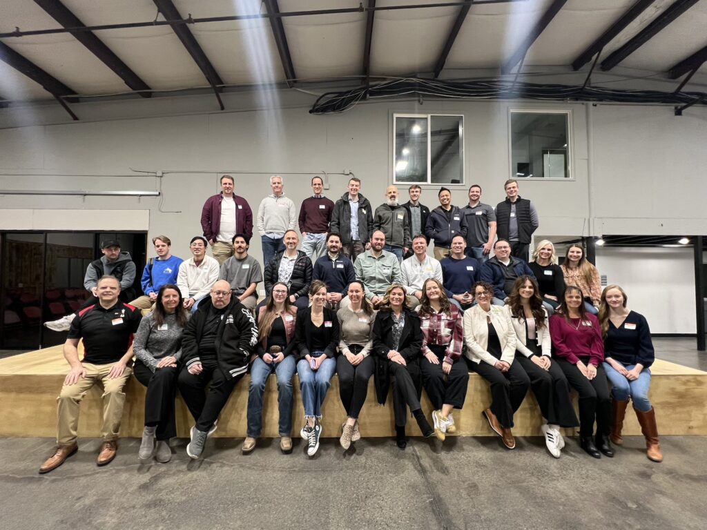 A large group of around 40 people posing together in a well-lit room. The group is smiling and dressed in casual to business-casual attire and standing/sitting on a wooden platform for a Utah Marketing Group meeting.