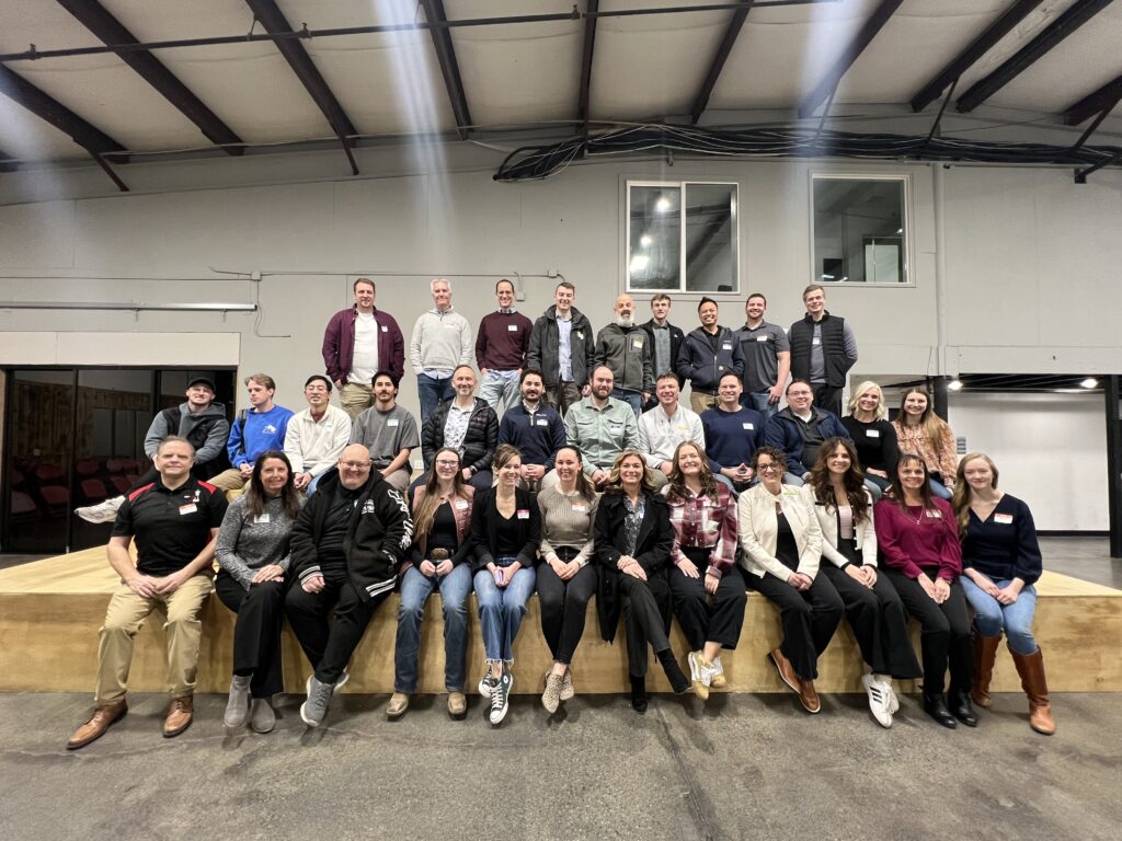 A large group of around 40 people posing together in a well-lit room. The group is smiling and dressed in casual to business-casual attire and standing/sitting on a wooden platform for a Utah Marketing Group meeting.