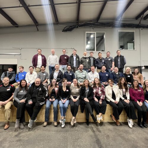 A large group of around 40 people posing together in a well-lit room. The group is smiling and dressed in casual to business-casual attire and standing/sitting on a wooden platform for a Utah Marketing Group meeting.