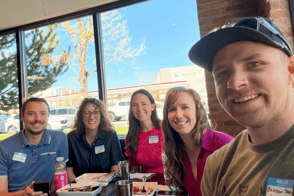 A small group of 5 people posing together in front of windows and around a dining table at Vessel Kitchen. The group is smiling and dressed in casual to business-casual attire and gathered for a Utah Marketing Group meeting.