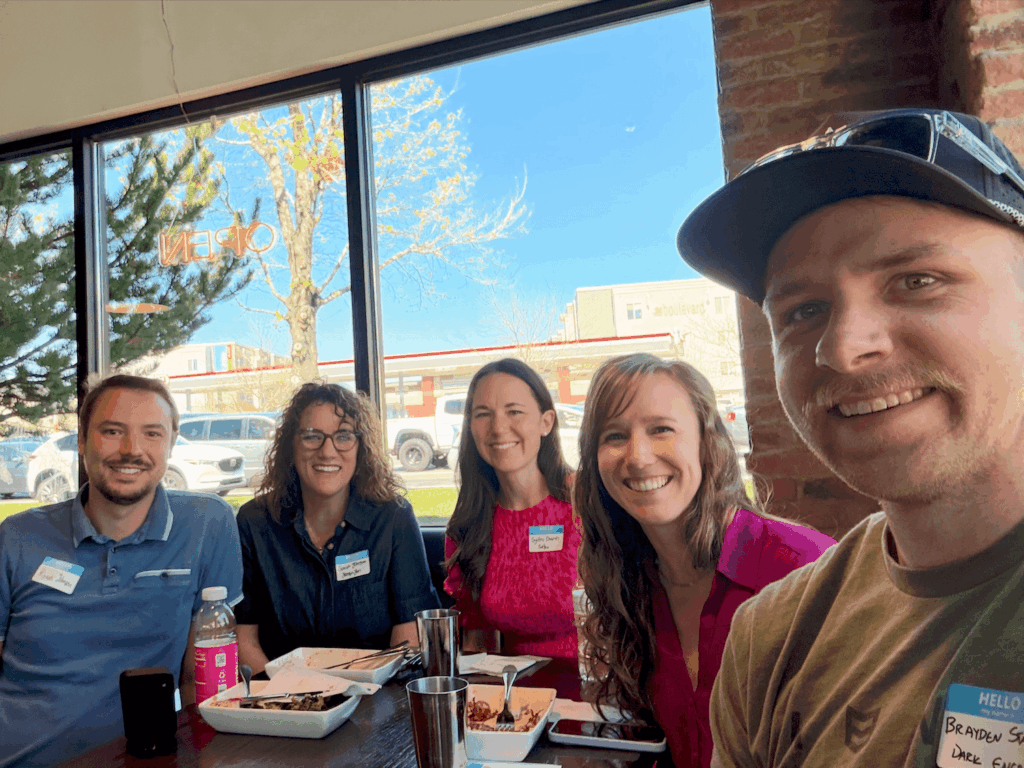 A small group of 5 people posing together in front of windows and around a dining table at Vessel Kitchen. The group is smiling and dressed in casual to business-casual attire and gathered for a Utah Marketing Group meeting.