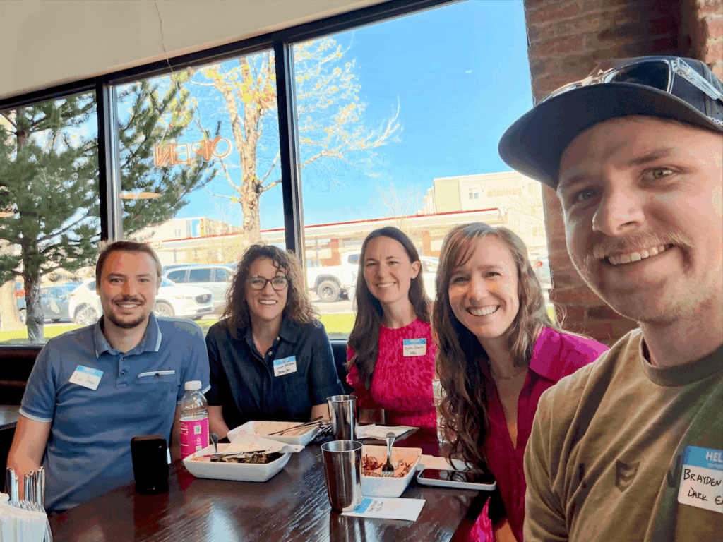 A small group of 5 people posing together in front of windows and around a dining table at Vessel Kitchen. The group is smiling and dressed in casual to business-casual attire and gathered for a Utah Marketing Group meeting.
