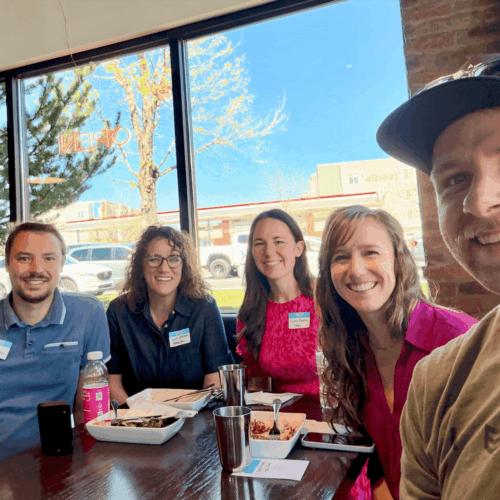 A small group of 5 people posing together in front of windows and around a dining table at Vessel Kitchen. The group is smiling and dressed in casual to business-casual attire and gathered for a Utah Marketing Group meeting.