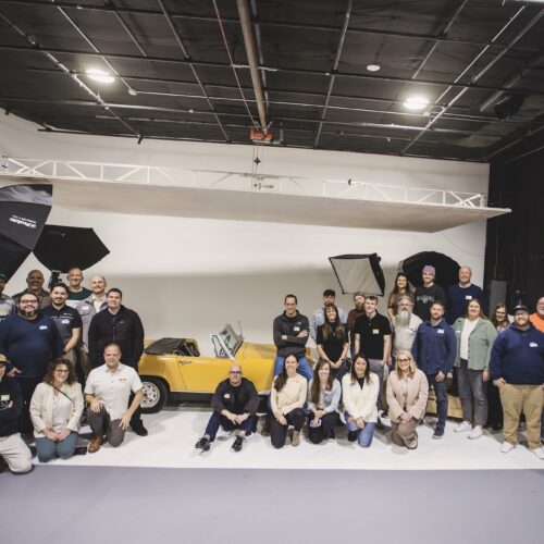 A group of about 40 people gathered for Utah Marketing Group, sitting in a photography studio, framing an old, yellow car.