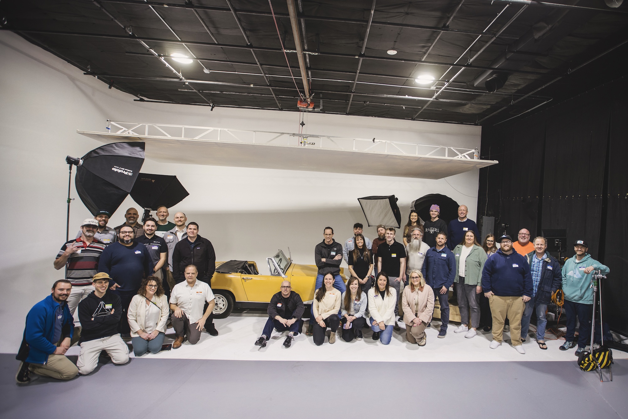 A group of about 40 people gathered for Utah Marketing Group, sitting in a photography studio, framing an old, yellow car.