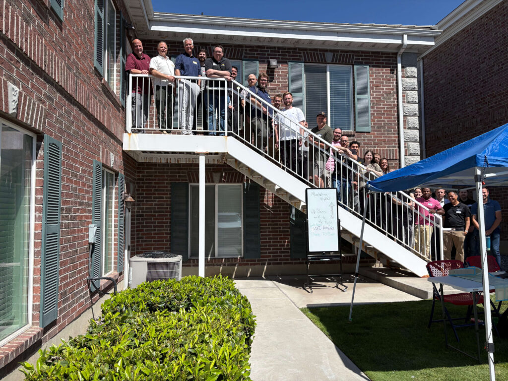 Attendees of a Utah Marketing Group networking event pose for a group photo on an outdoor staircase between two brick office buildings. Participants line the stairs and upper landing, smiling toward the camera, while a blue canopy tent and event sign are visible in the courtyard below on a sunny day.