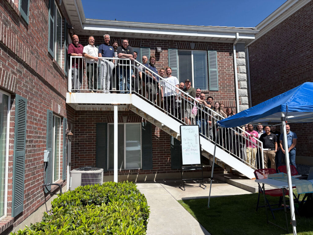 Attendees of a Utah Marketing Group networking event pose for a group photo on an outdoor staircase between two brick office buildings. Participants line the stairs and upper landing, smiling toward the camera, while a blue canopy tent and event sign are visible in the courtyard below on a sunny day.