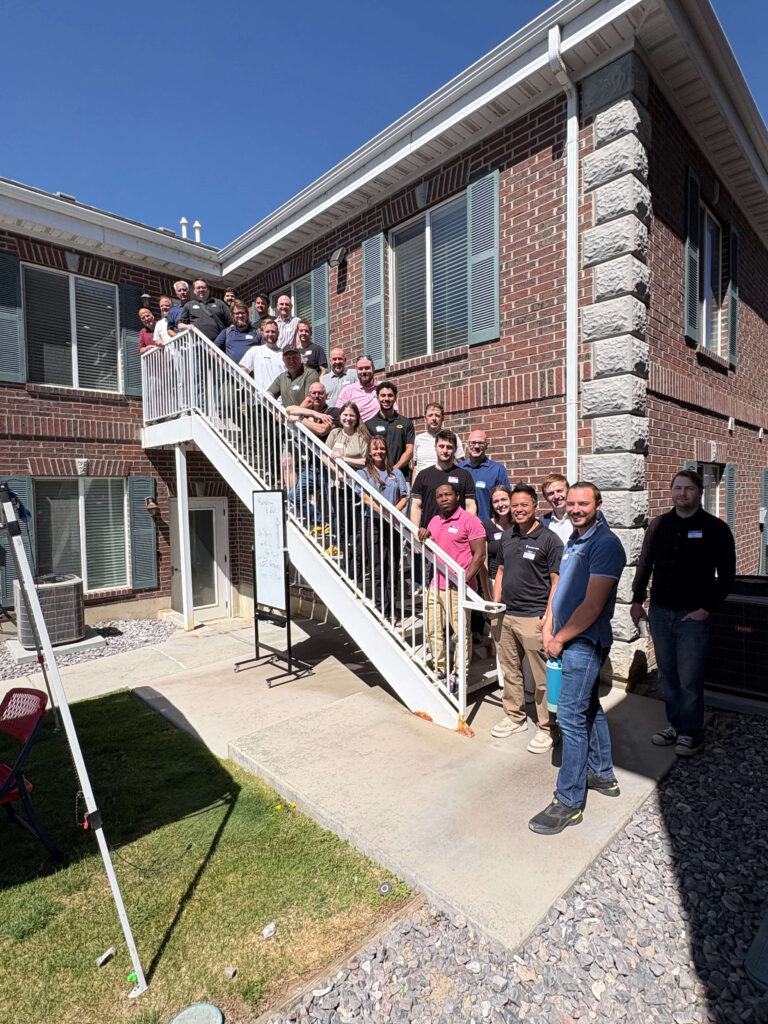 Attendees of a Utah Marketing Group networking event pose for a group photo on an outdoor staircase between two brick office buildings. Participants line the stairs and upper landing, smiling toward the camera, while a blue canopy tent and event sign are visible in the courtyard below on a sunny day.