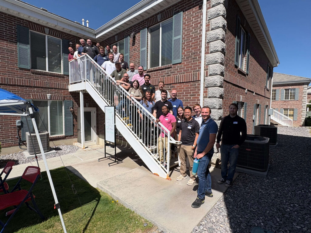 Attendees of a Utah Marketing Group networking event pose for a group photo on an outdoor staircase between two brick office buildings. Participants line the stairs and upper landing, smiling toward the camera, while a blue canopy tent and event sign are visible in the courtyard below on a sunny day.