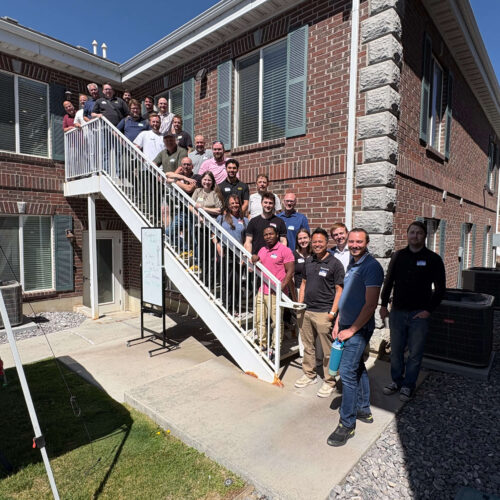 Attendees of a Utah Marketing Group networking event pose for a group photo on an outdoor staircase between two brick office buildings. Participants line the stairs and upper landing, smiling toward the camera, while a blue canopy tent and event sign are visible in the courtyard below on a sunny day.
