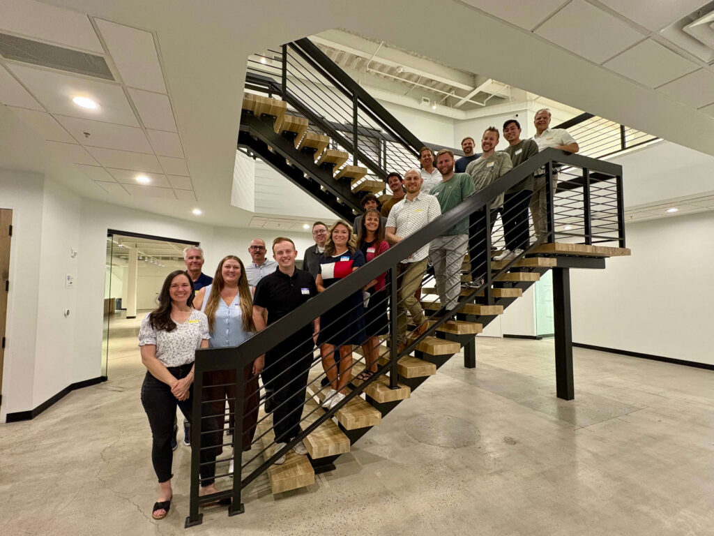 Attendees of a Utah Marketing Group networking event pose for a group photo on an indoor staircase in an office building. Participants line the stairs, smiling toward the camera.