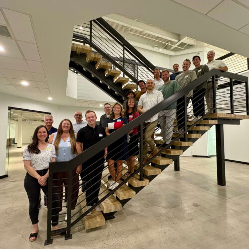 Attendees of a Utah Marketing Group networking event pose for a group photo on an indoor staircase in an office building. Participants line the stairs, smiling toward the camera.