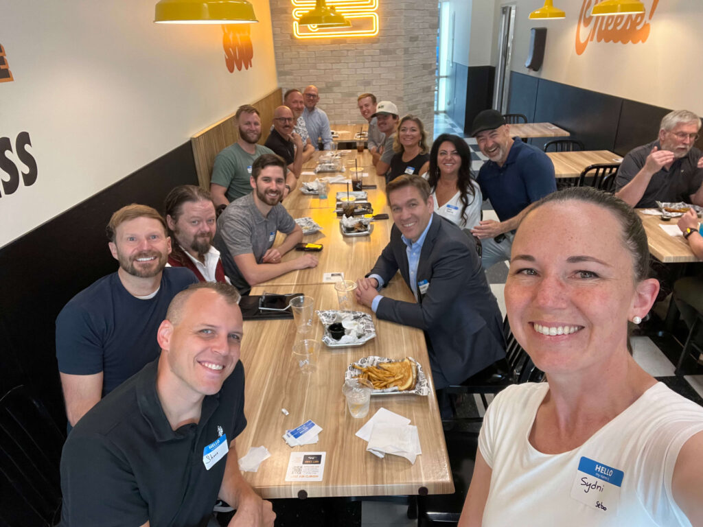 A group of 15 people posing together around a dining table at Melty restaurant. The group is smiling and dressed in casual to business-casual attire and gathered for a Utah Marketing Group meeting.