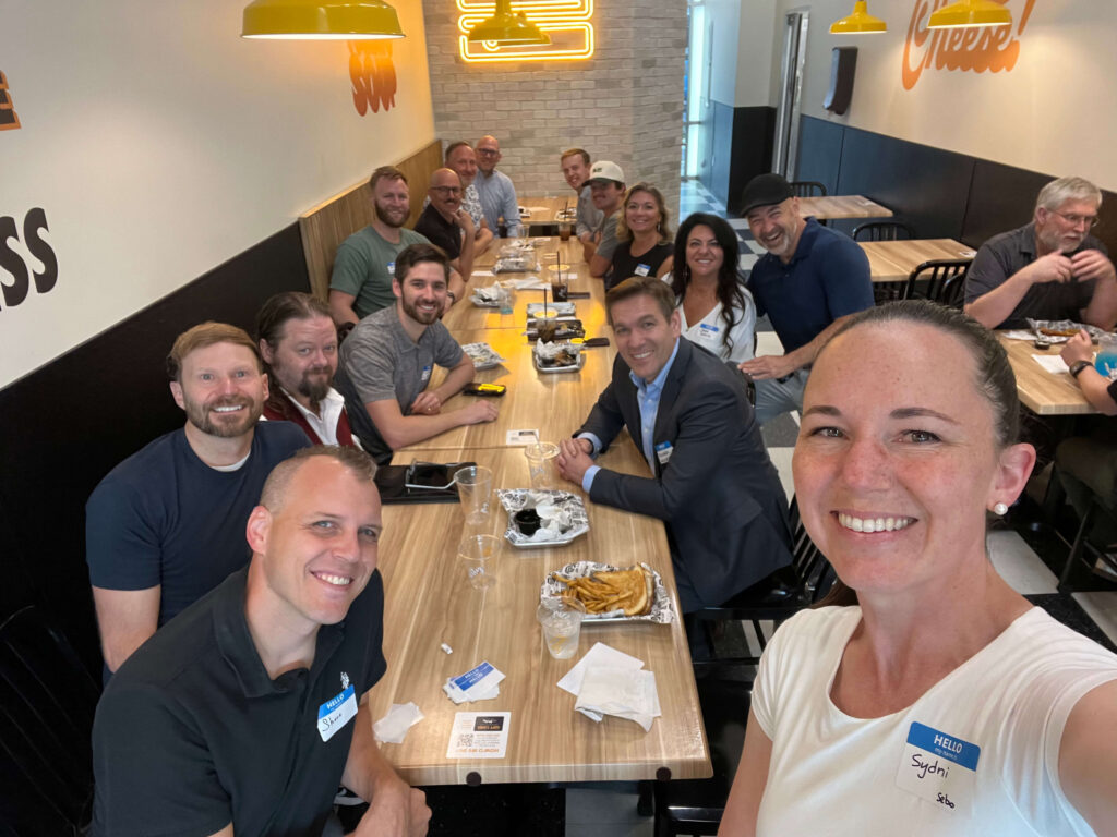A group of 15 people posing together around a dining table at Melty restaurant. The group is smiling and dressed in casual to business-casual attire and gathered for a Utah Marketing Group meeting.
