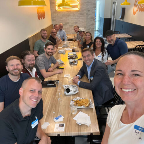 A group of 15 people posing together around a dining table at Melty restaurant. The group is smiling and dressed in casual to business-casual attire and gathered for a Utah Marketing Group meeting.