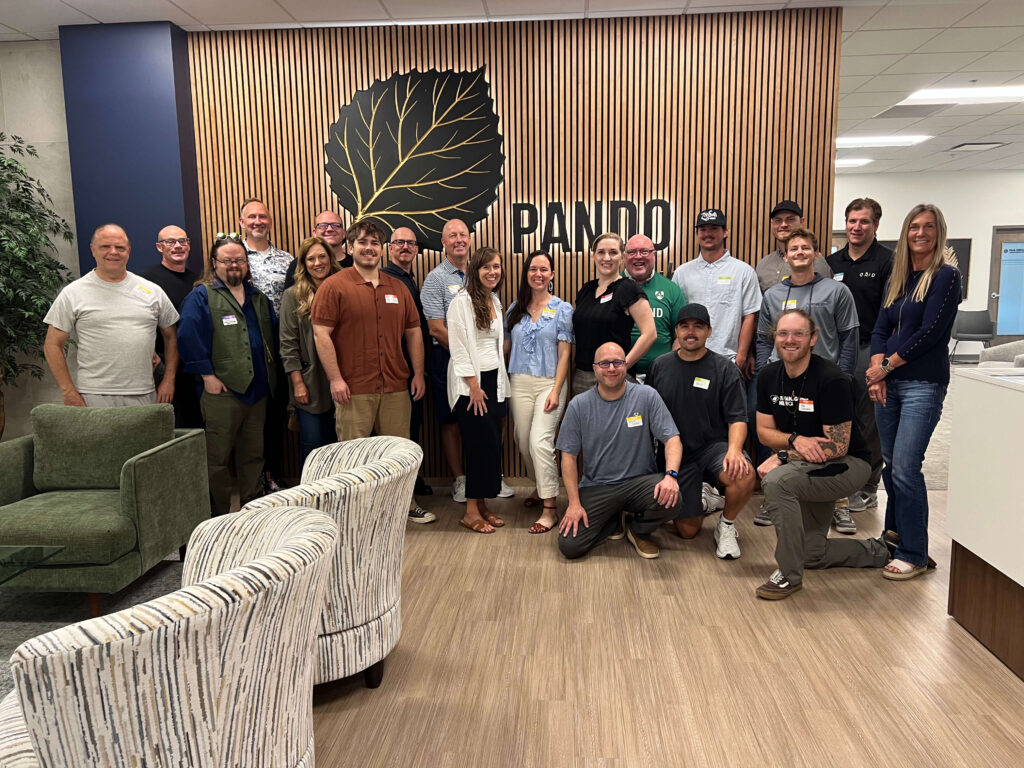 Group photo of Utah Marketing Group members gathered for a networking meeting at Pando Club, standing and kneeling together in front of the Pando logo and wood-paneled feature wall.