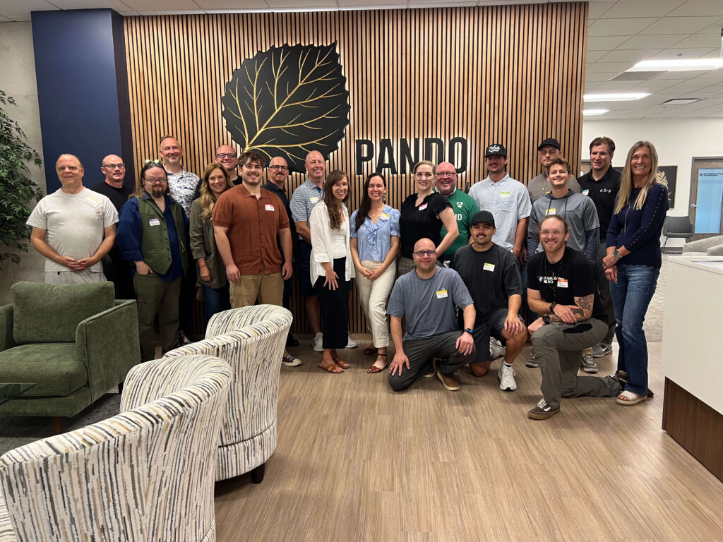 Group photo of Utah Marketing Group members gathered for a networking meeting at Pando Club, standing and kneeling together in front of the Pando logo and wood-paneled feature wall.