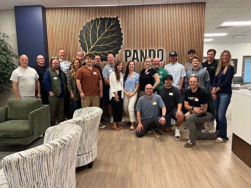 Group photo of Utah Marketing Group members gathered for a networking meeting at Pando Club, standing and kneeling together in front of the Pando logo and wood-paneled feature wall.